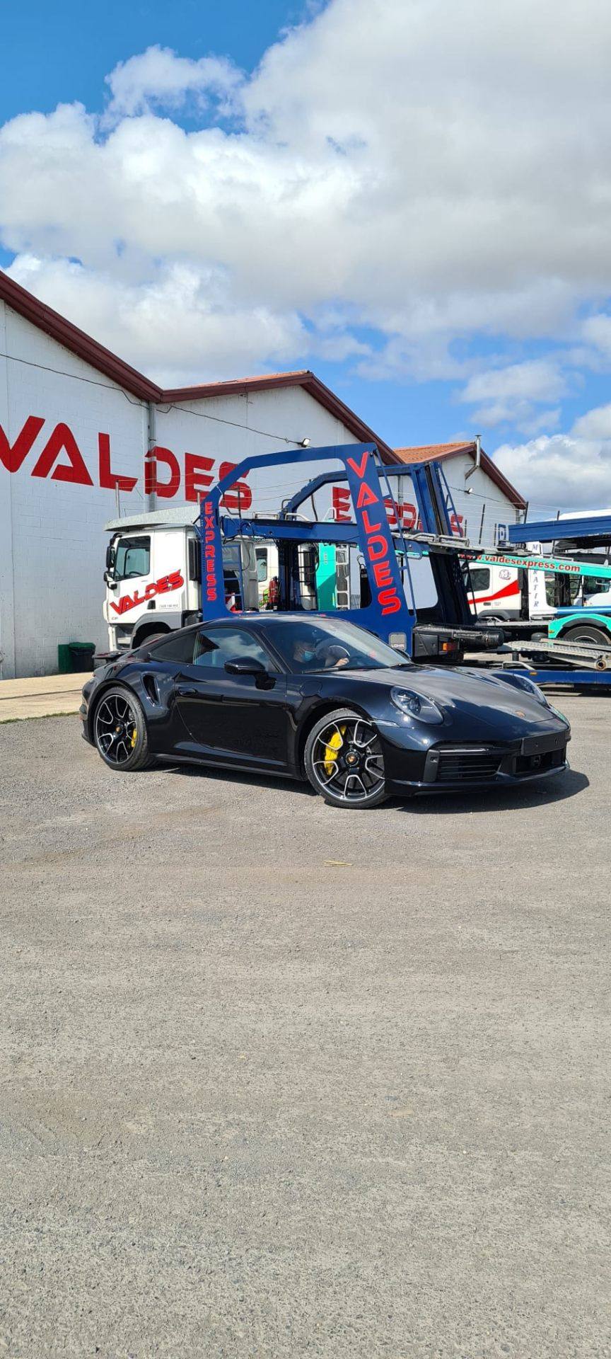 Un Porsche negro junto a la nave de Valdés Express, con un portavehículos detrás